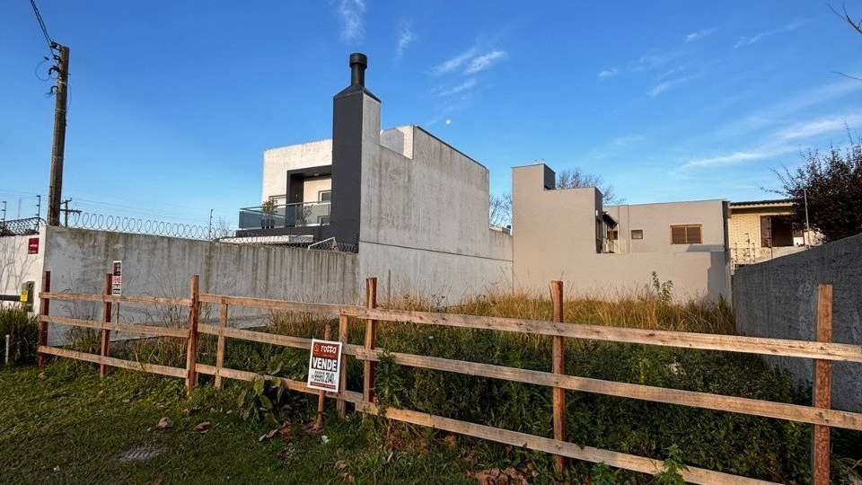 Terreno vazio com placa de venda cercado por muro e casas vizinhas ao fundo.