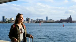 Mulher sorrindo com mochila e mala à beira-mar, cidade com prédios altos e igreja ao fundo.