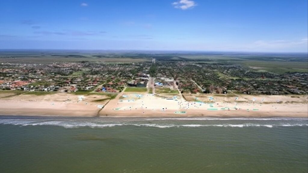 Vista aérea de cidade litorânea com faixa de areia larga, mar calmo e área urbana ao fundo.
