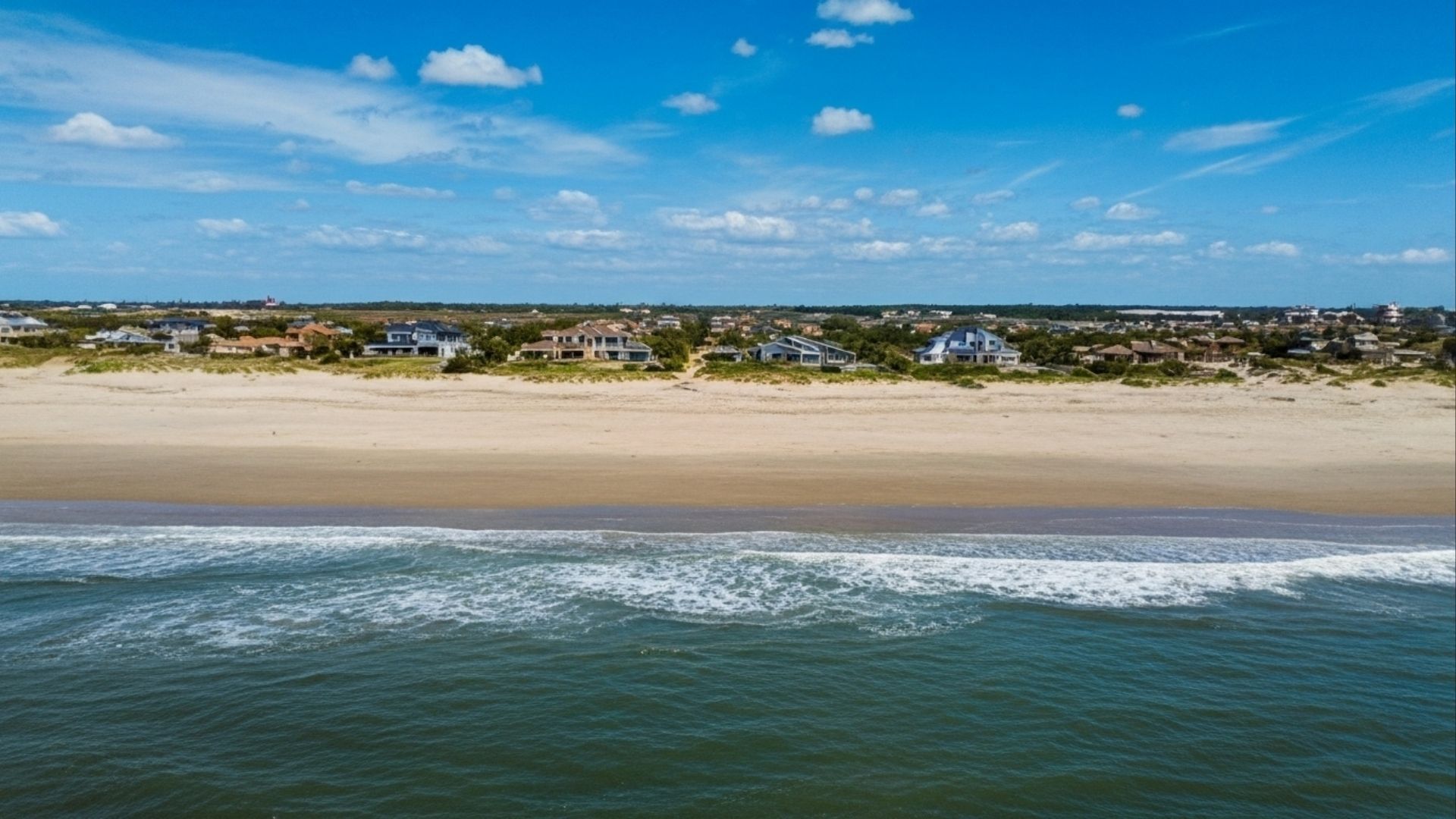 Vista aérea de casas de frente para o mar, separadas da água por ampla faixa de areia.