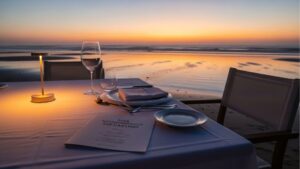 Mesa posta para jantar à beira-mar ao entardecer, com taças, talheres, guardanapo dobrado e cardápio com o título “Guia Gastronômico do Cassino”.