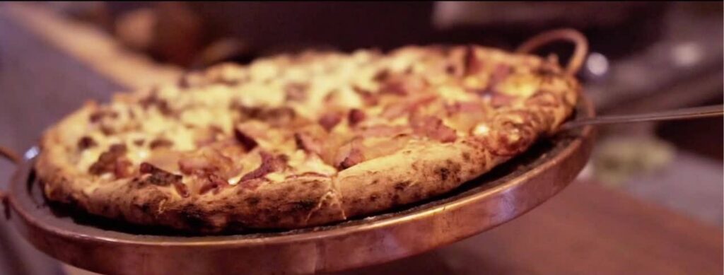 Pizza assada com borda grossa servida em bandeja metálica redonda, com cobertura de queijo, bacon e outros ingredientes.