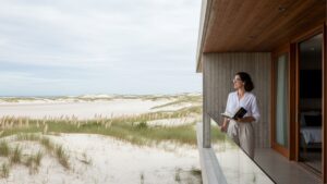 Mulher segurando livro em varanda de casa minimalista com vista para dunas de areia e oceano sob céu nublado.
