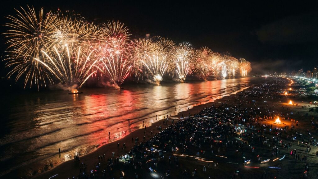 Vista aérea noturna de uma praia lotada com uma grandiosa queima de fogos de artifício acontecendo sobre o mar. O céu e a água estão iluminados pelos fogos em tons de dourado e avermelhado. Na extensa faixa de areia, milhares de pessoas acompanham o espetáculo e várias fogueiras estão acesas ao longo da costa.